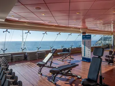 Ein heller Fitnessbereich an Bord der MSC Orchestra mit großen Fenstern zum Meer. Mehrere Hantelbänke und Hantelständer sind auf einem Holzboden arrangiert. Im Hintergrund ist das offene Meer zu sehen.