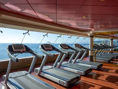 Moderner Fitnessraum an Bord der MSC Orchestra. Laufbänder stehen vor Fenstern mit Meerblick und Küstenlandschaft. Rote Decke, Holzfußboden.