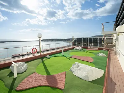 Eine Minigolf-Anlage an Deck der MSC Orchestra. Grüne Spielbahnen, rote Ziegelwege, weiße Fels-Hindernisse. Im Hintergrund: Meer, Hafen und Küstenlinie unter blauem Himmel. Keine Personen.