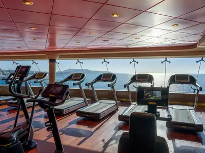 Das moderne Fitnessstudio der MSC Orchestra zeigt Laufbänder und Ellipsentrainer mit Panoramablick auf das Meer und die Küste. Der Raum ist hell beleuchtet und menschenleer.