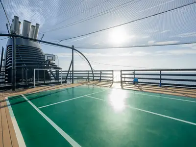 Sportplatz auf Oberdeck der MSC Orchestra. Grüne Fläche mit Basketballkorb und Fußballtor. Hintergrund: Schiffsschornstein, Meer, blauer Himmel, umgeben von Schutznetz. Sonne glänzt auf dem Feld.