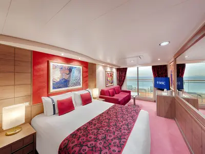 Eine helle Premium Suite Aurea an Bord der MSC Orchestra. Das Zimmer hat ein Doppelbett mit roten Kissen, einen roten Teppich und eine rote Couch. Ein Balkon mit Meerblick ist durch Glasschiebetüren sichtbar.