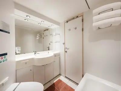 Helles Badezimmer einer Premium Suite Aurea an Bord der MSC Orchestra. Es zeigt ein Waschbecken mit Armatur, einen großen Spiegel, ein WC und eine Badewanne. Gerollte Handtücher hängen an der Wand.