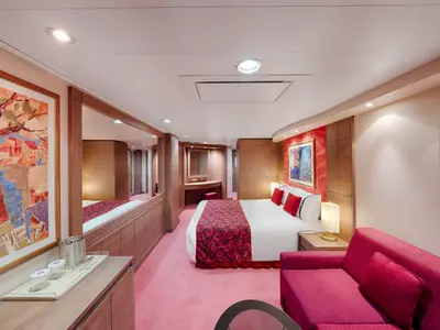 Eine Premium Suite Aurea an Bord der MSC Orchestra. Der Raum zeigt ein großes Doppelbett mit roter Tagesdecke, ein rotes Sofa, einen Schminktisch und eine helle Holzvertäfelung.