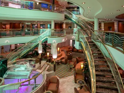 Ansicht des eleganten Atriums an Bord der MSC Poesia. Eine mehrstufige, geschwungene Treppe mit goldfarbenen Geländern und gemusterter Glasarchitektur führt durch den Raum. Im unteren Bereich befindet sich ein Lounge-Bereich mit Sesseln und Sofas sowie ein transparentes Klavier.