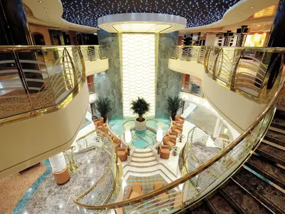 Das Atrium der MSC Poesia von oben. Eine beleuchtete Wand, ein Wasserbecken mit Palmen und orangefarbene Sessel. Elegante goldene Treppengeländer führen zu den oberen Etagen.