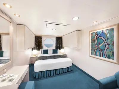 Eine gemütliche Deluxe Aussenkabine an Bord der MSC Poesia. Ein Doppelbett mit blauen Kissen und Bettrock dominiert die Mitte, zum runden Bullaugenfenster mit Meerblick ausgerichtet. Rechts hängt ein großes, abstraktes Kunstwerk über einem blauen Sessel. Links ist ein Schminktisch mit Spiegel sichtbar. Der Boden ist mit blauem Teppich ausgelegt.
