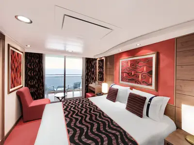 Eine helle und modern eingerichtete Balkonkabine an Bord der MSC Poesia mit einem Doppelbett, rotem Sessel und einem Balkon mit Meerblick. Rote Wände und dunkle Holzmöbel dominieren den Raum.