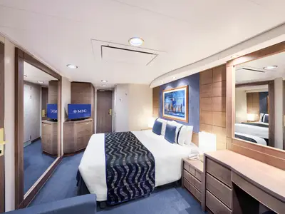 Balkonkabine an Bord der MSC Poesia mit Doppelbett, blauer Tagesdecke, dunklen Holzvertäfelungen, Flachbildfernseher und Schminkbereich mit Spiegel.
