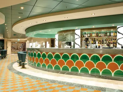 Eine moderne Bar an Bord der MSC Poesia mit einem langen Thekenbereich. Die Theke ist mit beleuchteten orange-grünen Schuppenmustern verziert und hat eine Marmorplatte. Dahinter stehen Flaschen mit Spirituosen auf Regalen. Grüne Barhocker sind vor der Theke platziert. Der Raum ist hell beleuchtet.