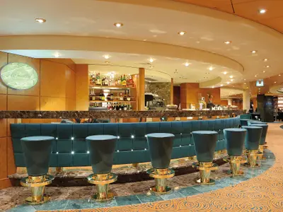 Bar Smeraldo an Bord der MSC Poesia. Grüne Barhocker mit goldenen Details vor einer Marmortheke. Beleuchtetes Logo und viele Flaschen im Hintergrund.
