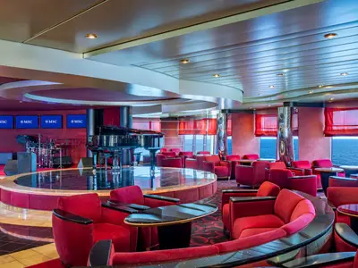 Innenansicht der Pigalle Lounge an Bord der MSC Poesia. Eine Bühne mit glänzend schwarzem Flügel und Schlagzeug steht im Vordergrund. Rote Sessel und kleine Tische bieten Sitzgelegenheiten entlang großer Fenster mit Blick auf das offene Meer.