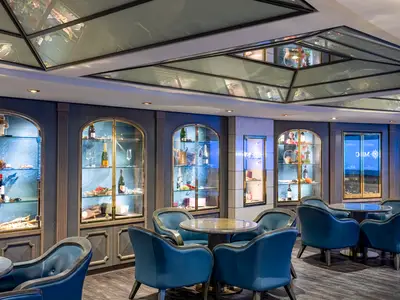 Die Il Grappolo d'Oro Bar-Lounge an Bord der MSC Poesia. Blaue Sessel und Tische stehen vor beleuchteten Vitrinen mit Weinen und Champagner. Die Decke ist mit reflektierenden Glaselementen versehen. Ohne Personen.