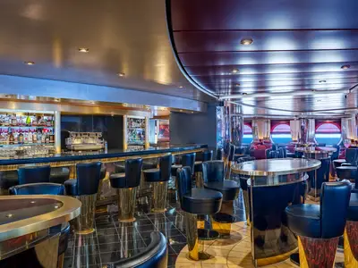 Innenansicht der Pigalle Lounge an Bord der MSC Poesia, mit einer voll ausgestatteten Bar, schwarzen Barhockern mit goldenen Füßen und runden Tischen. Große Bullaugen bieten Blick auf das Meer.