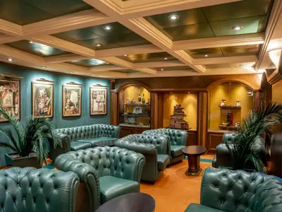 Elegante Lounge an Bord der MSC Poesia. Dunkelgrüne Chesterfield-Sofas und Sessel, petrolfarbene Wände mit Kunst, beleuchtete Holz-Vitrinen. Orangefarbener Teppich.