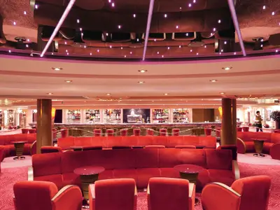 Die Bar Dei Poeti an Bord der MSC Poesia zeigt eine luxuriöse Lounge mit plüschigen roten Sofas und Sesseln. Ein langer Bartresen mit reich gefüllten Regalen ist im Hintergrund sichtbar, beleuchtet von warmem Licht. Zwei Crewmitglieder sind zu sehen. Die Decke ist mit violetten Lichtpunkten versehen.