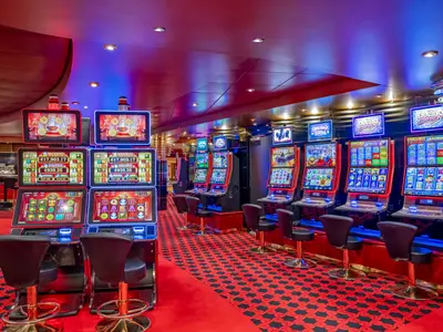 Das lebhafte Casino Royal an Bord der MSC Poesia zeigt Reihen von modernen Slotmaschinen mit bunten Bildschirmen. Der Raum ist mit roten Teppichen und Akzentlichtern in Rot und Blau ausgestattet, leere Hocker stehen vor den Automaten.