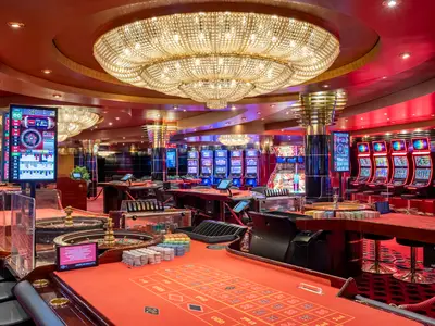 Elegantes Casino Royal der MSC Poesia. Rote Roulette-Tische, Jetons und Bildschirme im Vordergrund. Dahinter Reihen von Spielautomaten und glitzernde Kronleuchter.