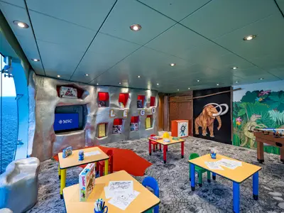 Ein heller Kinderclub I Dinosauri an Bord der MSC Poesia mit grossen Fenstern zum Meer. Es gibt bunte Tische, Stühle, Dinosaurier-Wandmalereien und eine Lego-Wand.