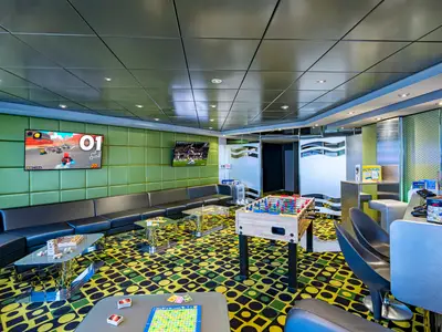 Ein lebhafter Spielbereich an Bord der MSC Poesia mit grünen Wänden, einem Foosball-Tisch, Videospielen auf Bildschirmen und bequemen Sitzgelegenheiten zum Entspannen und Spielen. Brettspiele liegen auf glänzenden Tischen bereit.