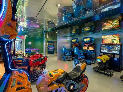 Lebhafte Indoor-Spielhalle an Bord der MSC Poesia. Viele Arcade-Automaten: Rennsimulatoren, Schießspiele, Greifautomat. Helle Neonlichter, reflektierende Decke. Ohne Personen.