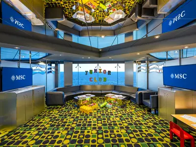 Der bunte Junior Club der MSC Poesia zeigt einen grossen Spielbereich mit bequemen Sofas, Tischen voller Spiele und Bausteinen. Grosse Fenster bieten Meerblick, Bildschirme zeigen das MSC Logo.