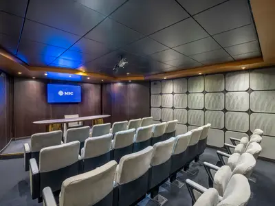 Blick in das Poesia Meeting Center an Bord der MSC Poesia. Der Raum ist mit eleganten Stoffsitzen in Reihen, einem grossen Bildschirm mit MSC-Logo und einer Holzvertäfelung ausgestattet. Deckenlichter tauchen den Raum in ein warmes Licht, während der Bildschirm blau leuchtet.