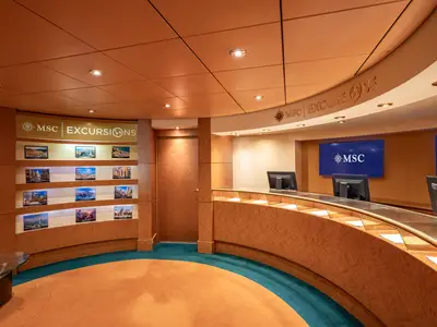 Ein heller, moderner Landausflugsbereich an Bord der MSC Poesia mit einem geschwungenen Holzthekenbereich, Bildschirmen mit dem MSC-Logo und einer Wand voller Reisefotos.