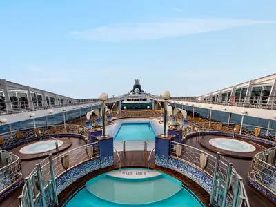Die Poollandschaft mit Hauptpool und mehreren Whirlpools an Deck der MSC Poesia. Liegestühle, eine grosse Leinwand und der Schornstein sind zu sehen. Blauer Himmel.