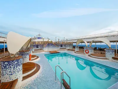Der weitläufige Poolbereich an Deck der MSC Poesia bietet mehrere Becken und Liegeflächen. Blaue Mosaikfliesen und Holz verzieren die Gestaltung. Im Hintergrund ist das offene Meer sichtbar.