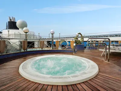 Ein sprudelnder Whirlpool auf dem Teakholz-Sonnendeck der MSC Poesia, mit Blick auf die Schiffsaufbauten, eine Skulptur und das weite Meer unter blauem Himmel.