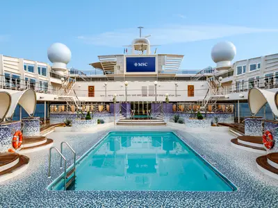 Blick auf das Hauptdeck der MSC Poesia mit einem grossen, rechteckigen Pool in der Mitte, umgeben von blau-weissen Mosaikfliesen und Holzdecks. Im Hintergrund ist eine grosse Videoleinwand mit dem MSC-Logo zu sehen.