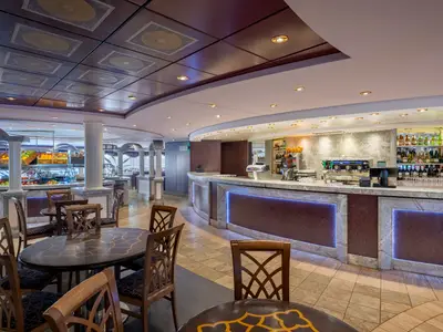 Ein heller und geräumiger Buffetbereich an Bord der MSC Poesia mit frischen Früchten, Marmortresen und einem voll ausgestatteten Barbereich mit blauer LED-Beleuchtung. Mehrere Tische und Stühle sind im Vordergrund zu sehen.
