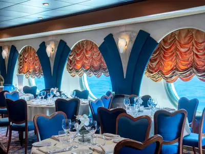 Blick in das elegant eingerichtete Restaurant Le Fontane an Bord der MSC Poesia, mit zahlreichen gedeckten Tischen, blauen Stühlen und runden Fenstern, die den Blick auf das Meer freigeben. Die Vorhänge sind gold-orange, die Wände und Stühle blau.