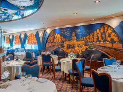 Innenansicht des eleganten Restaurants 'Le Fontane' an Bord der MSC Poesia. Blaue Stühle und Sofas, weiß gedeckte Tische. Ein großes Wandgemälde zeigt eine Fontäne und goldene Bäume. Durch Fenster ist das blaue Meer zu sehen.
