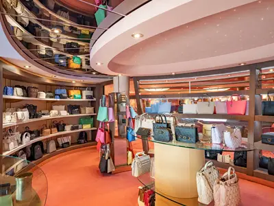 Ein elegant beleuchteter Accessoires-Shop an Bord der MSC Poesia zeigt Regale und Glasvitrinen voller Designer-Handtaschen in verschiedenen Farben und Stilen. Der Boden ist mit einem roten Teppich ausgelegt und die Decke ist gewölbt mit integrierten Lichtern.