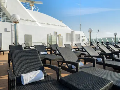 Sun deck on MSC Poesia with rows of modern dark wicker sun loungers, each with a rolled white towel. The ship's white superstructure and blue sky are in the background.