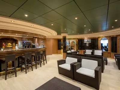 Elegante Bar-Lounge an Bord der MSC Poesia. Eine Holztheke mit Barhockern, frischem Obst und Getränken ist zu sehen. Bequeme Korbsessel mit weißen Kissen stehen im Raum.
