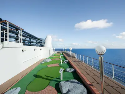 Der Minigolfplatz an Deck der MSC Poesia unter blauem Himmel. Grüne Bahnen, rote Ziegelumrandungen, silberne Pfosten und das blaue Meer sind zu sehen. Die Sonne wirft lange Schatten. Keine Personen.