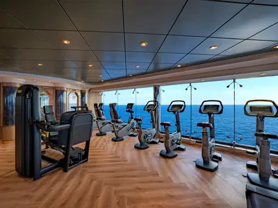 Fitnessraum an Bord der MSC Poesia mit modernen Technogym-Geräten. Panoramafenster bieten weiten Blick auf das Meer. Helle Holzoptik und gute Beleuchtung.