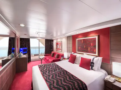 Geräumige Suite an Bord der MSC Poesia mit einem großen Bett, rotem Teppich, rotem Sofa und Balkon mit Blick auf das offene Meer. Warme Beleuchtung und Kunstwerke an den Wänden prägen das Ambiente.