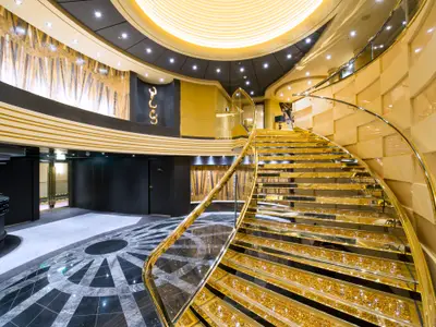Ein luxuriöses Atrium an Bord der MSC Preziosa mit einer breiten, geschwungenen Treppe. Die Stufen sind mit goldenen Swarovski-Kristallen besetzt und die Wände sind in Goldtönen gehalten. Die Szene ist hell erleuchtet.