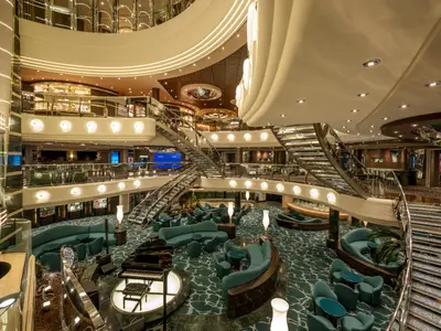 Blick von oben auf das elegante Atrium der MSC Preziosa mit mehreren Ebenen, verglasten Treppen, einer Pianobar, hellen Sitzbereichen in Petrol und Geschäften.