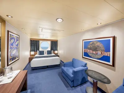 Eine moderne Aussenkabine der MSC Preziosa mit Doppelbett, Bullauge und blauem Teppichboden. Ein Sessel und Kunstwerke sind ebenfalls zu sehen.