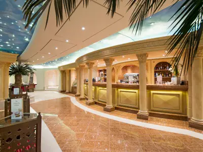 Blick in die elegante Phoenician Plaza Bar an Bord der MSC Preziosa. Eine geschwungene Theke mit Säulen dominiert den Raum, darüber eine Decke im Sternenhimmel-Design. Im Vordergrund sind Tische und Stühle zu sehen, im Hintergrund eine Palme und weitere Sitzbereiche. Der Boden ist gefliest.