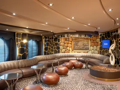 Safari Lounge an Bord der MSC Preziosa. Geschwungene Sitzbank, Leopardenmuster-Teppich, braune Hocker, Safari-Wände, MSC-Bildschirm, Leoparden-Kunstwerk.