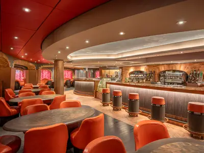 Ein stilvoller Bar- und Loungebereich an Bord der MSC Preziosa mit rötlichen Tönen, bequemen orangefarbenen Sesseln, Holztischen und einer langen Bar mit Flaschenregalen und Hockern.