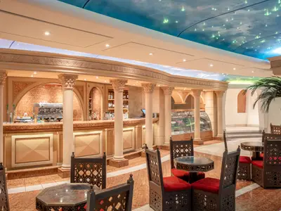 Die elegante Bar-Lounge Phoenician Plaza an Bord der MSC Preziosa zeigt eine lange Theke, verzierte Saeulen, Tische und Stuehle. Die Decke ist als Sternenhimmel mit Wolken gestaltet, ein Palmbaum steht im Hintergrund.
