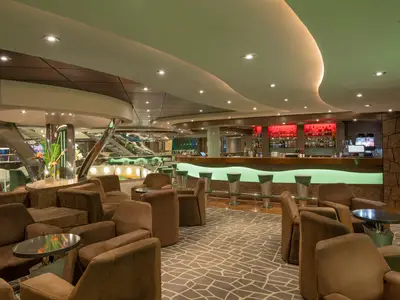 Die Diamond Bar der MSC Preziosa. Moderne Lounge: braune Sessel, Tische, grün leuchtende Bar. Rote Regalbeleuchtung. Glänzende Treppen im Hintergrund. Stilvolles Ambiente.