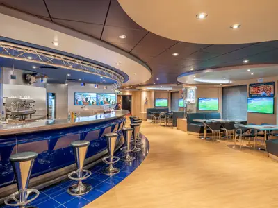 Eine moderne Sports Bar an Bord der MSC Preziosa mit einer langen blauen Bar, hohen Barhockern und mehreren Bildschirmen, die Sport zeigen. Im Hintergrund bequeme Lounge-Sitzbereiche.